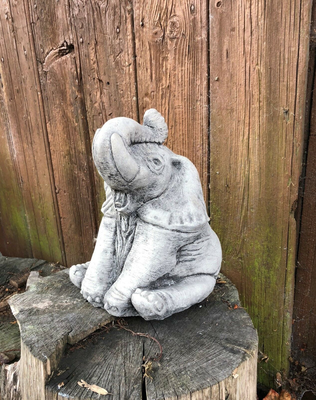 Cute stone baby elephant ornament with trunk raised, placed on a wooden stump, perfect for whimsical garden or porch decor.