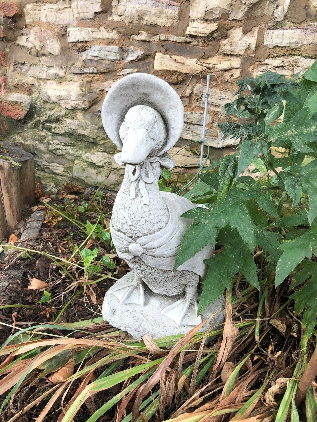 Handmade stone duck statue with bonnet, placed among plants and dry grass. Ideal for outdoor garden or porch decoration.
