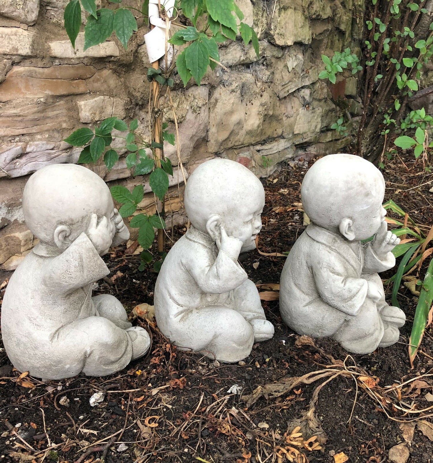 Side view of See, Hear, Speak No Evil Baby Monk Ornaments in stone finish, aligned in garden soil for decorative landscaping.