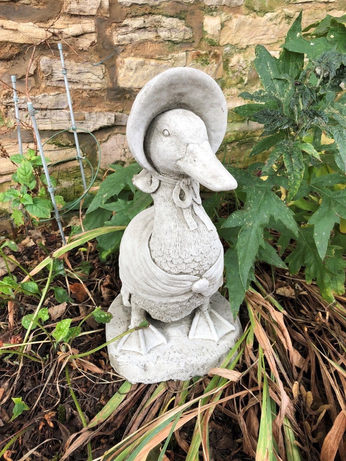 Whimsical duck statue in bonnet and shawl, crafted from stone and set in garden foliage. Great for lawn or balcony decoration.
