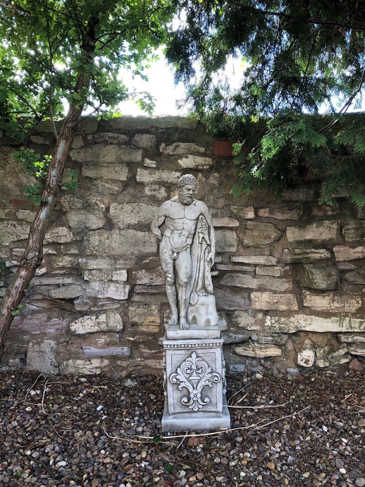 Centered view of Hercules stone statue in garden against stone wall, standing on ornate limestone pedestal.