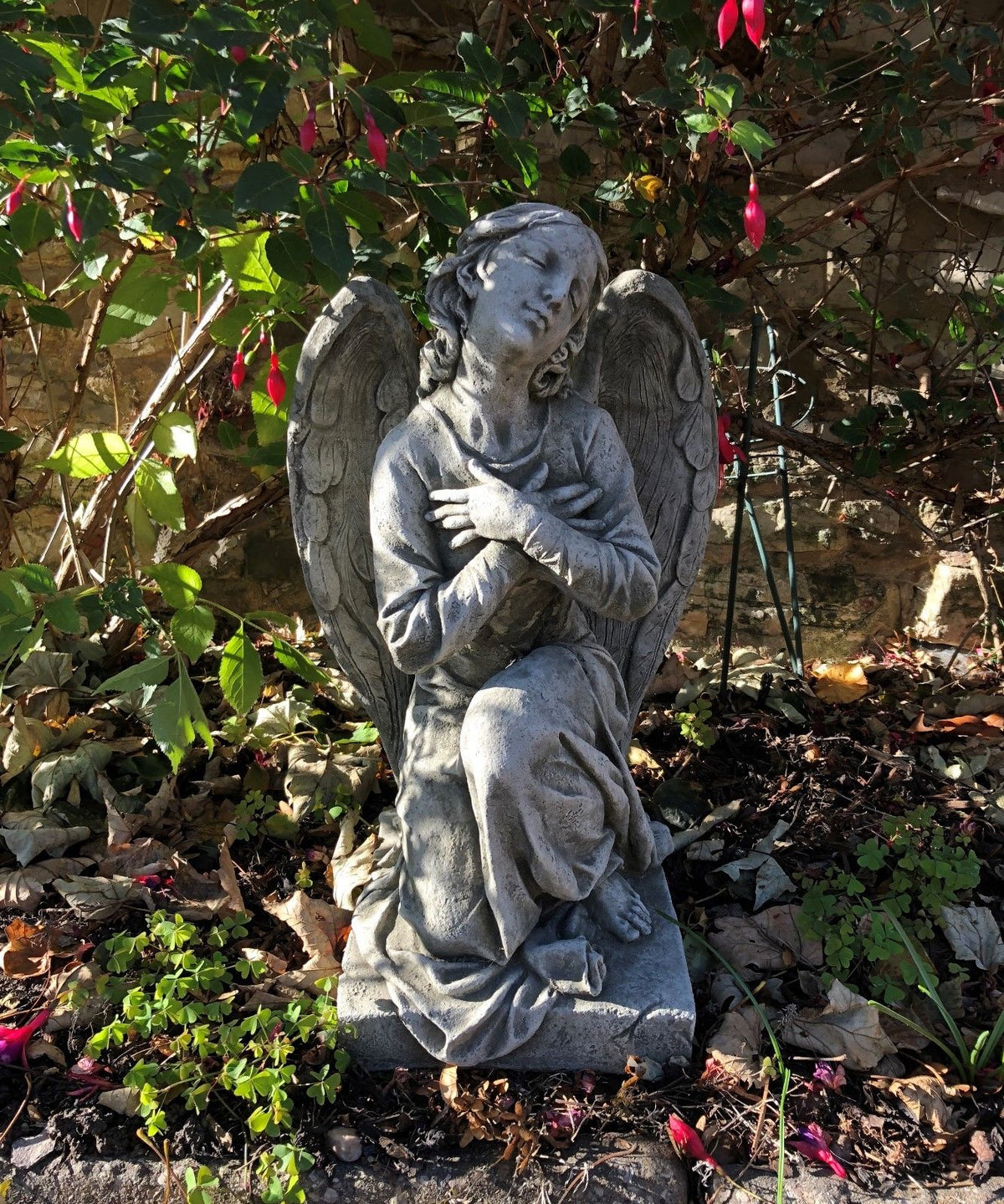 Angel Garden Statue - Stone Kneeling Arms Crossed placed in a flower bed, featuring detailed wings and serene expression for outdoor decor.
