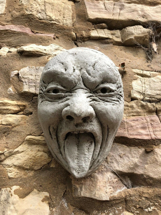 Stone Tongue Out Face Plaque mounted on rustic stone wall, adds cheeky charm to garden décor. Durable limestone for outdoor use.