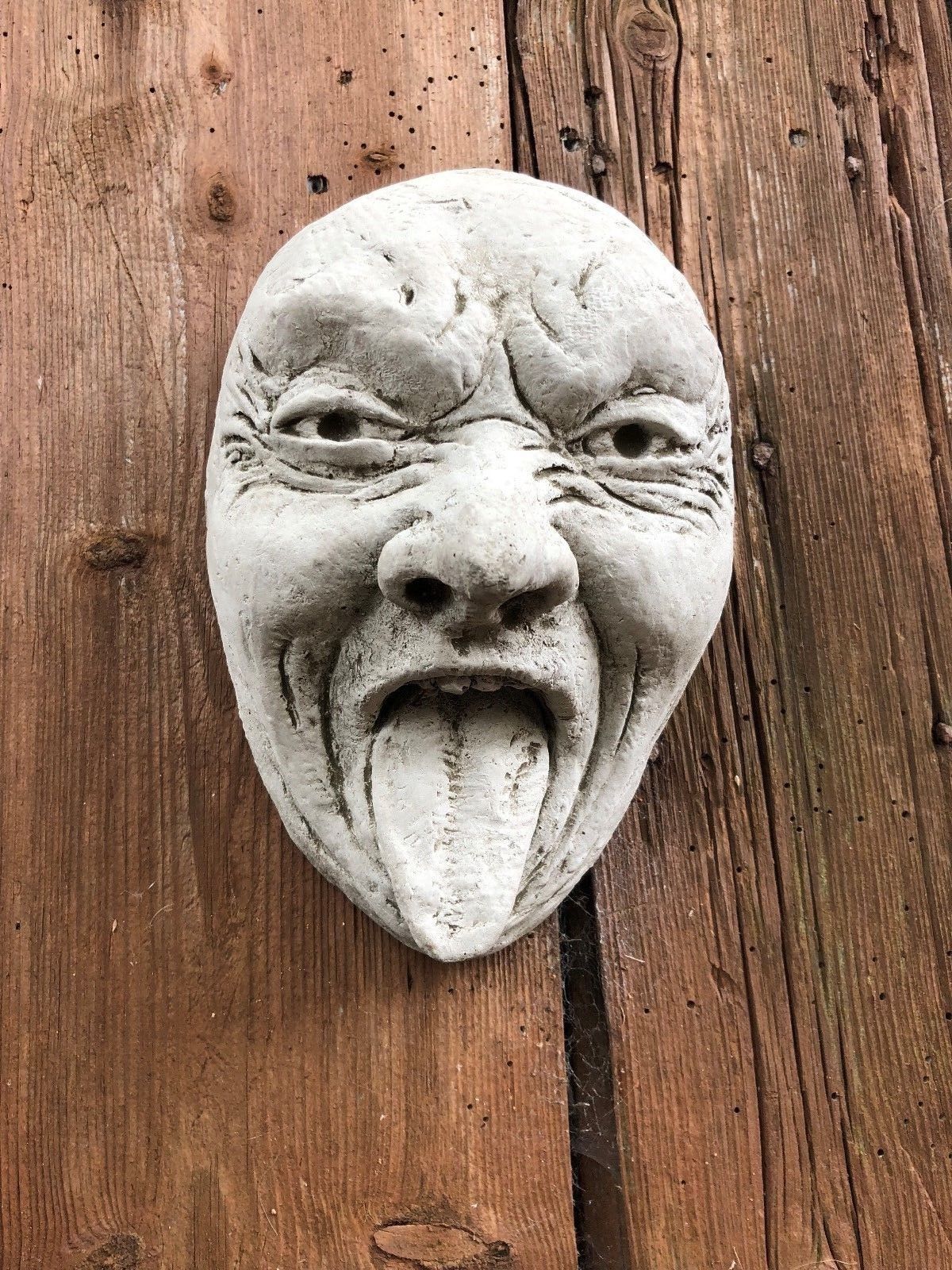 Stone Tongue Out Face Plaque on wooden wall, perfect for quirky garden decoration. Weather-resistant limestone sculpture.