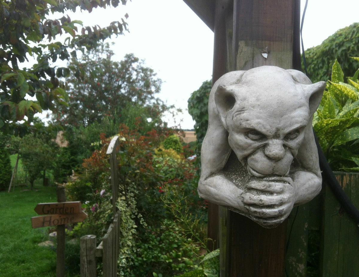 Stone Sculpture - Gargoyle Garden Ornament mounted on wooden post in lush garden. Gothic wall decor with antique limestone finish.