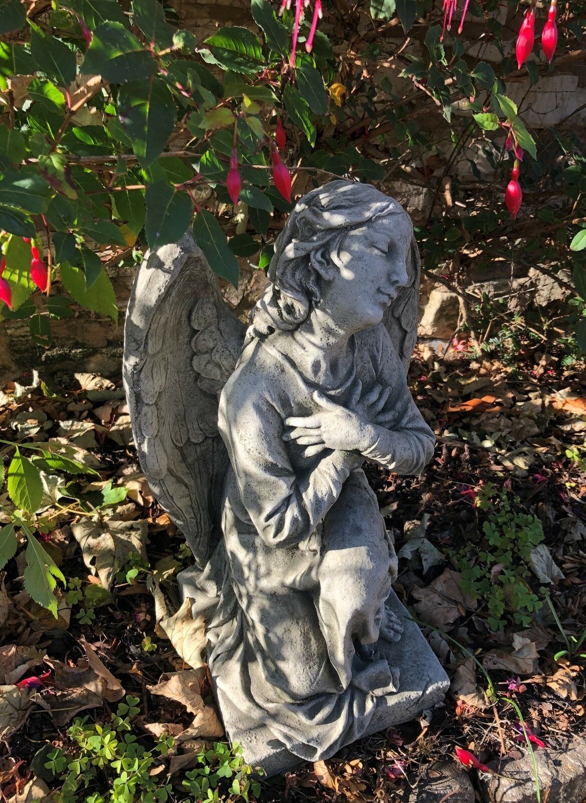 Side angle of Angel Garden Statue - Stone Kneeling Arms Crossed nestled in garden foliage, showcasing graceful posture and fine sculptural detail.