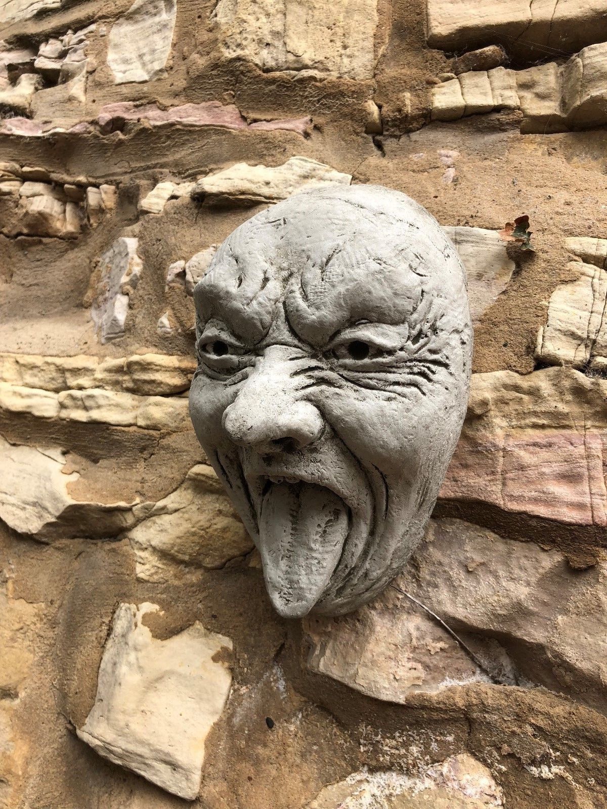 Side view of Stone Tongue Out Face Plaque on stone wall, showcasing detailed expression. Ideal for outdoor garden humor.