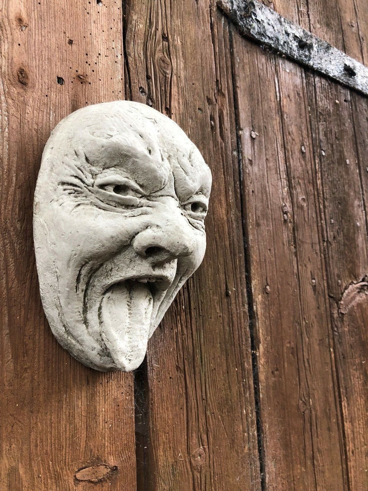 Angled view of Stone Tongue Out Face Plaque on wooden surface, highlighting textured limestone finish. Great for patios or fences.