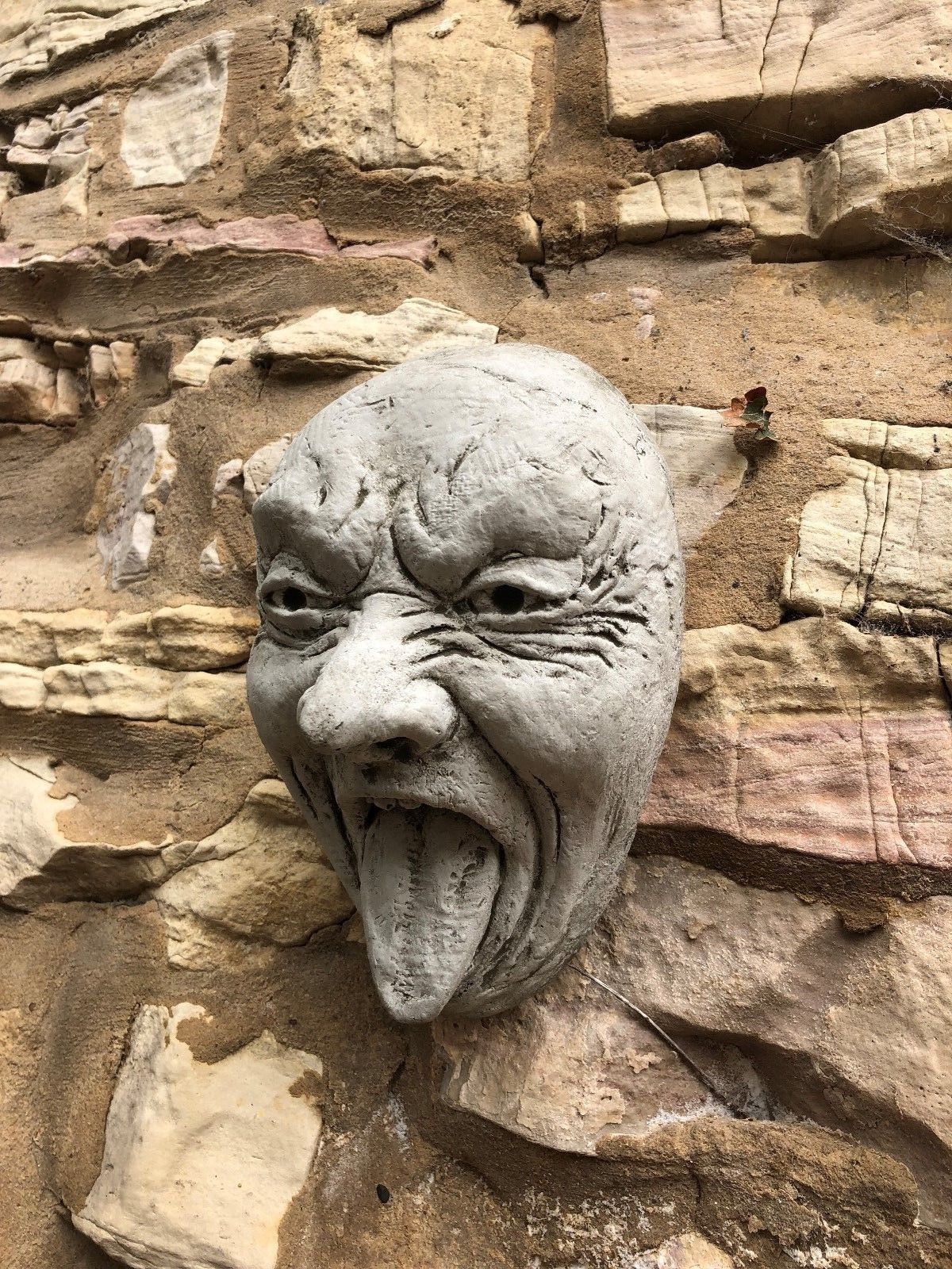 Stone Tongue Out Face Plaque affixed to rustic stone wall, features expressive design. A fun addition to garden ornaments.