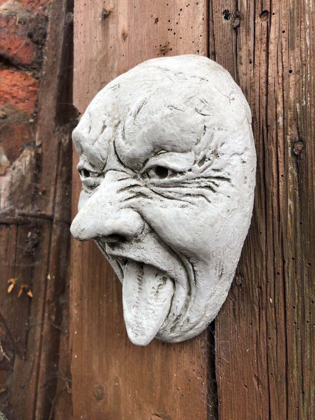 Close-up side view of Stone Tongue Out Face Plaque on wood, emphasizing handcrafted detail. Unique garden wall decoration.