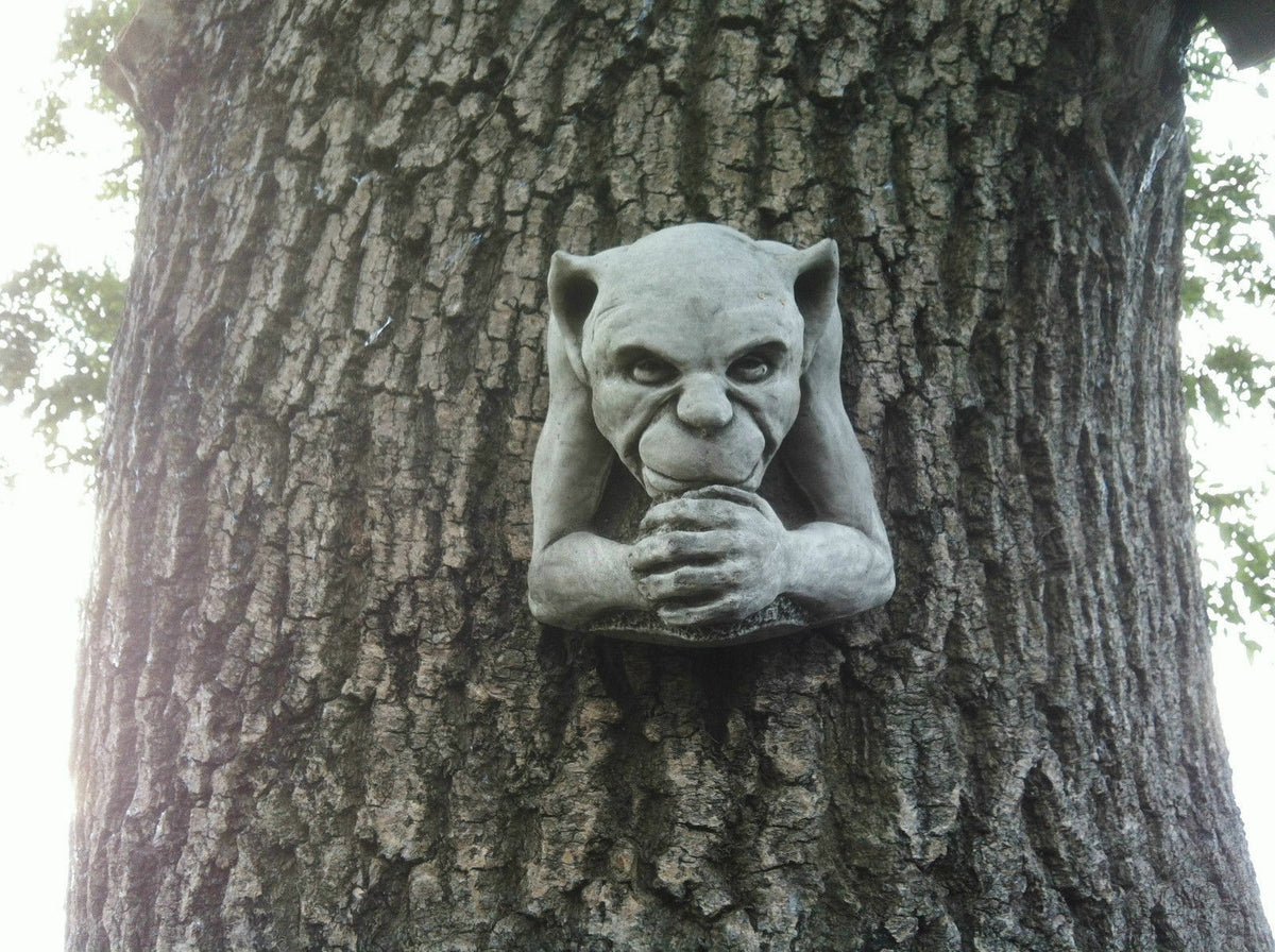 Gargoyle Garden Ornament mounted on tree trunk. Weather-resistant stone sculpture adds gothic charm to outdoor decor.