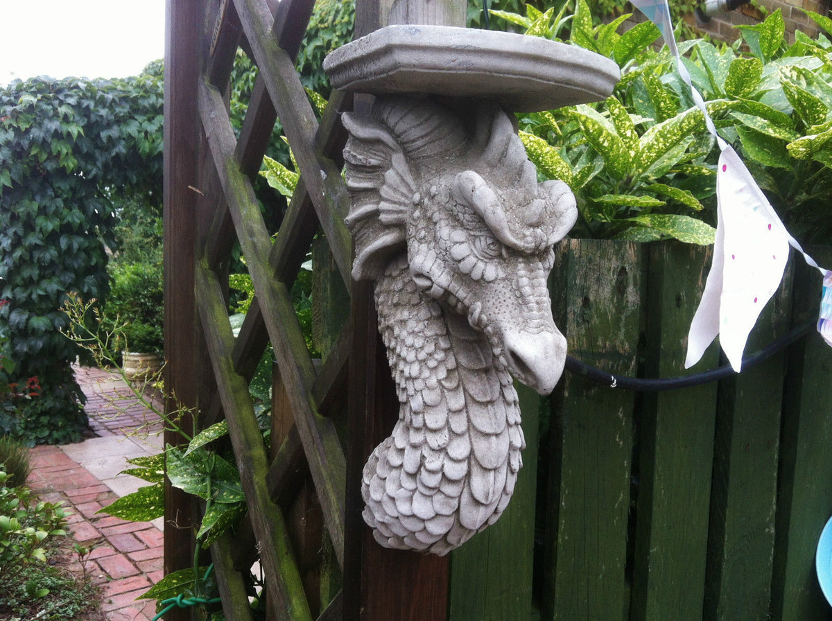 Close-up of Stone Dragon Wall Shelf on wooden post surrounded by greenery. Weather-resistant garden ornament for outdoor decoration.