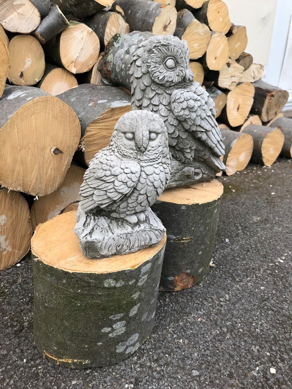 Mother and baby stone owl ornaments displayed on wooden logs. Durable limestone statues for garden or patio decoration.