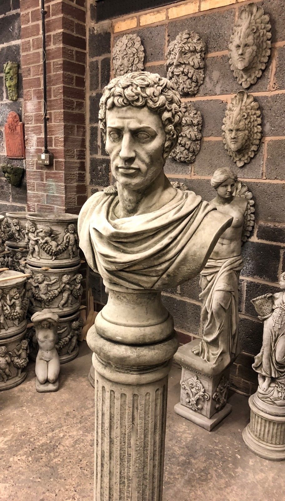 Angled view of Stone Marc Anthony Bust Statue with detailed toga and Roman styling, set on a limestone column for outdoor garden display.