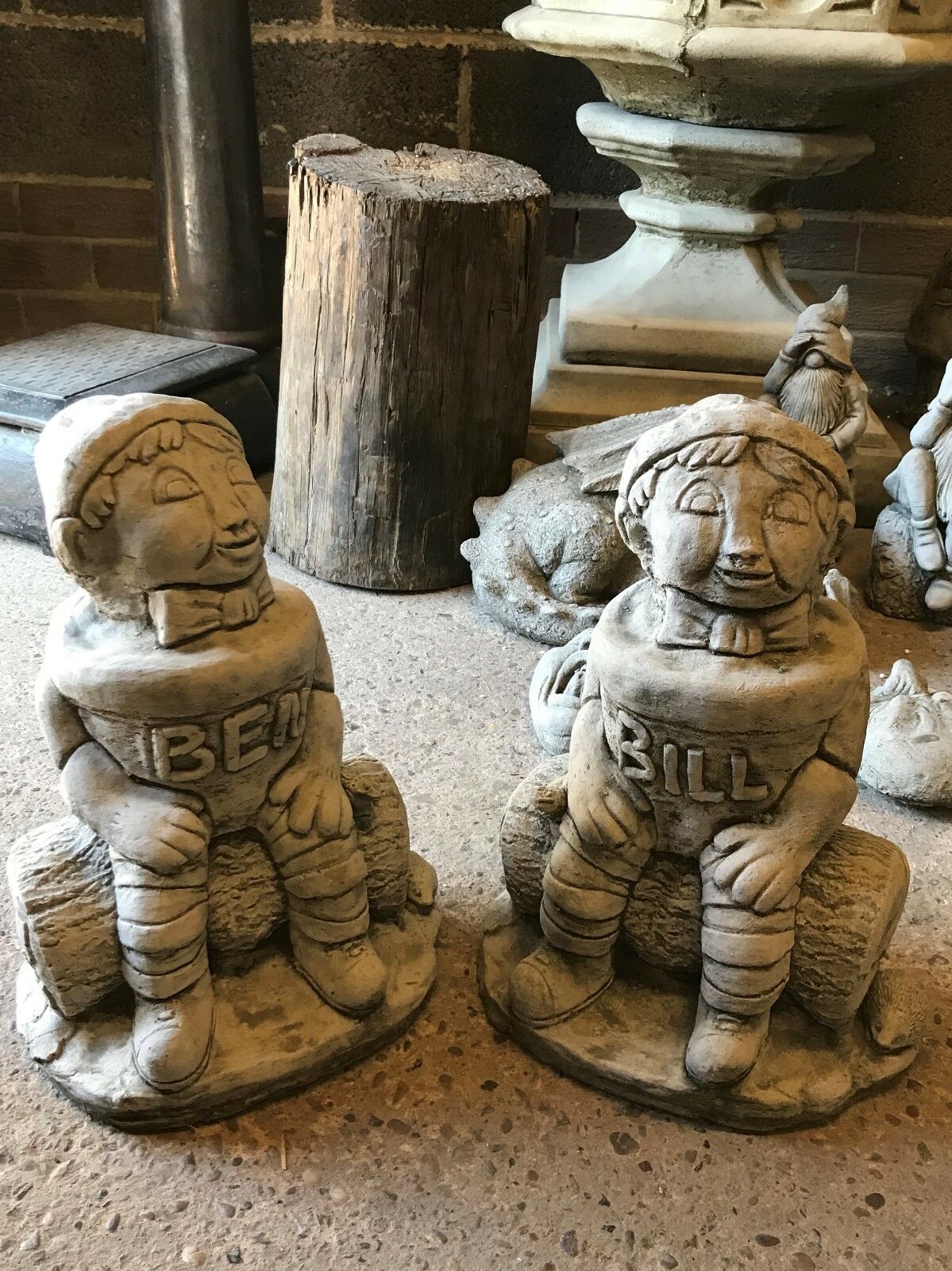Pair of Stone Bill & Ben statues with antique finish, seated on logs, perfect as durable and decorative garden ornaments.