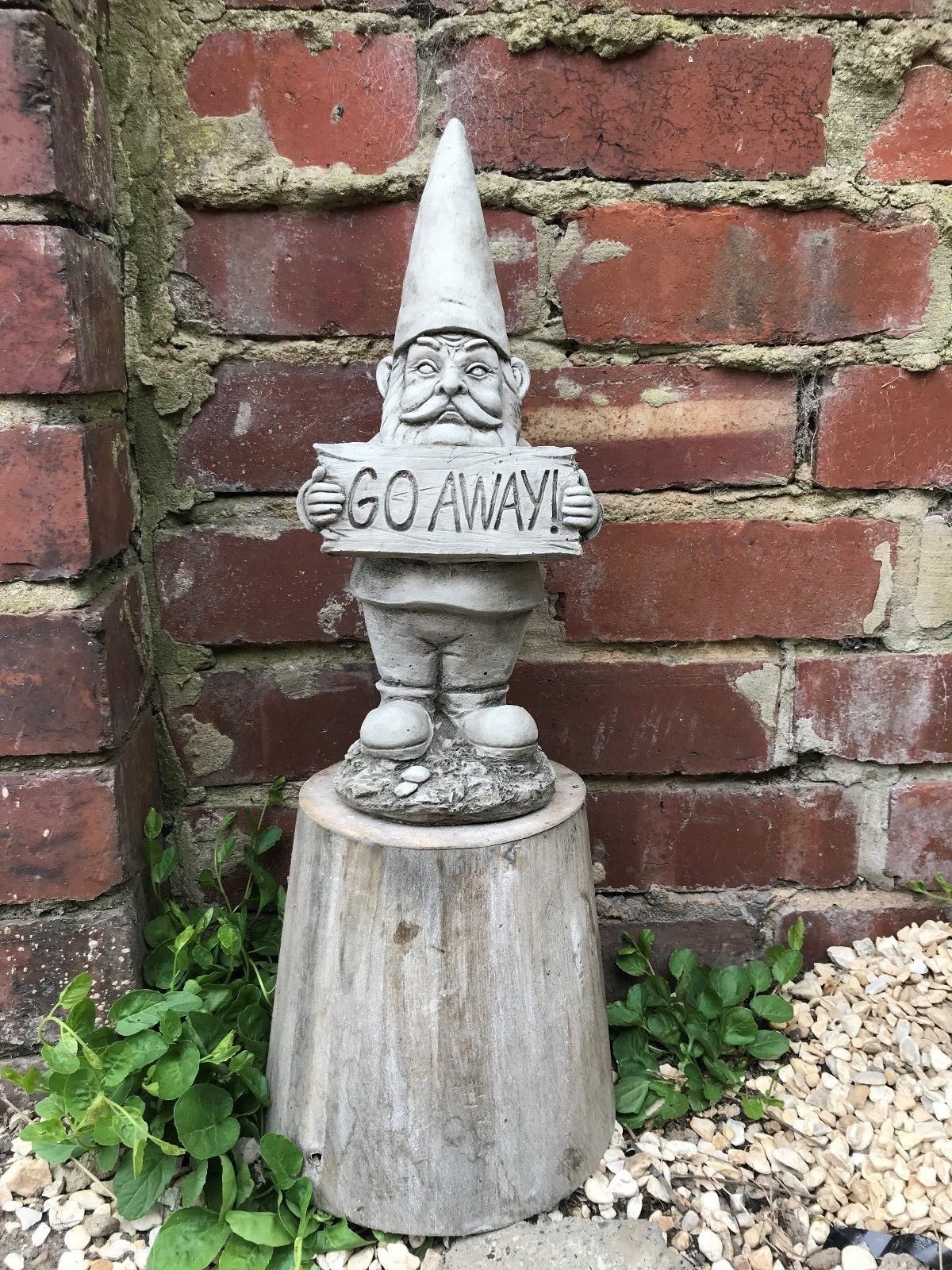 Grumpy Garden Gnome 'Go Away' ornament made from durable limestone, placed on a wooden stump against a brick wall in a garden setting.