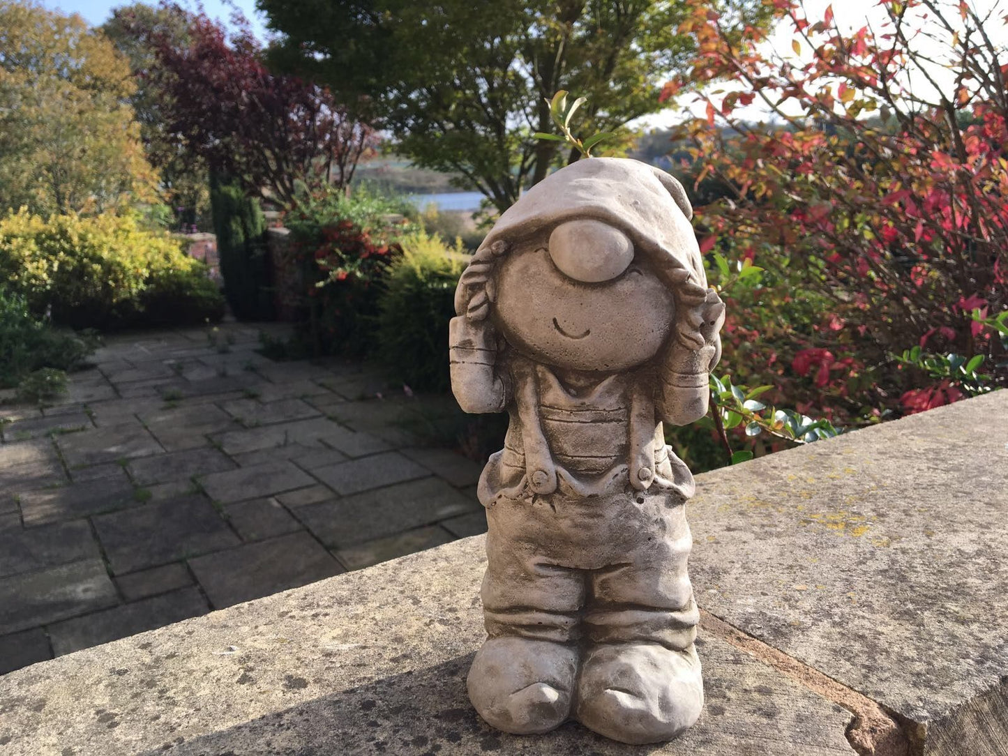 Stone Boy Garden Ornament in Dungarees standing on a stone ledge with a scenic garden view. Ideal for outdoor decor in patios or flower beds.