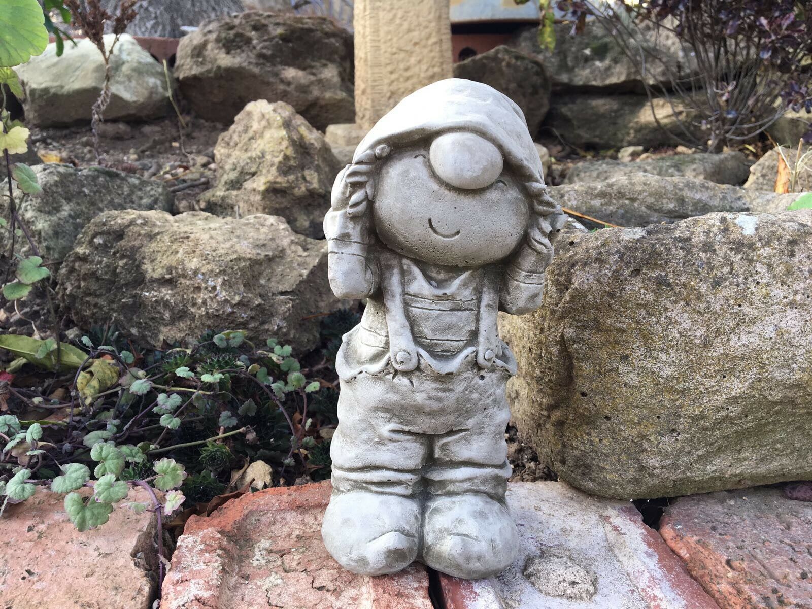 Charming Stone Boy Garden Ornament in Dungarees placed among rustic rocks. Perfect for enhancing garden aesthetics with a whimsical touch.
