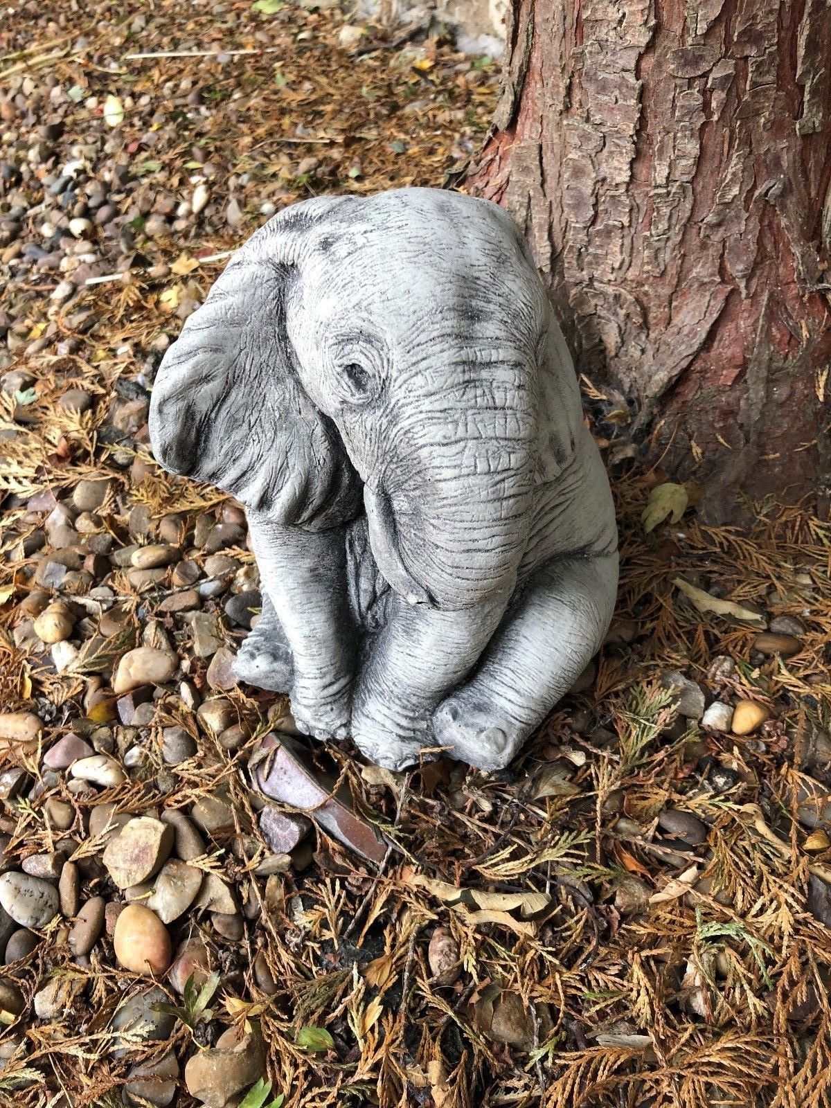 Grey stone baby elephant sculpture with lifelike features, placed on pebbled ground for decorative garden or patio enhancement.