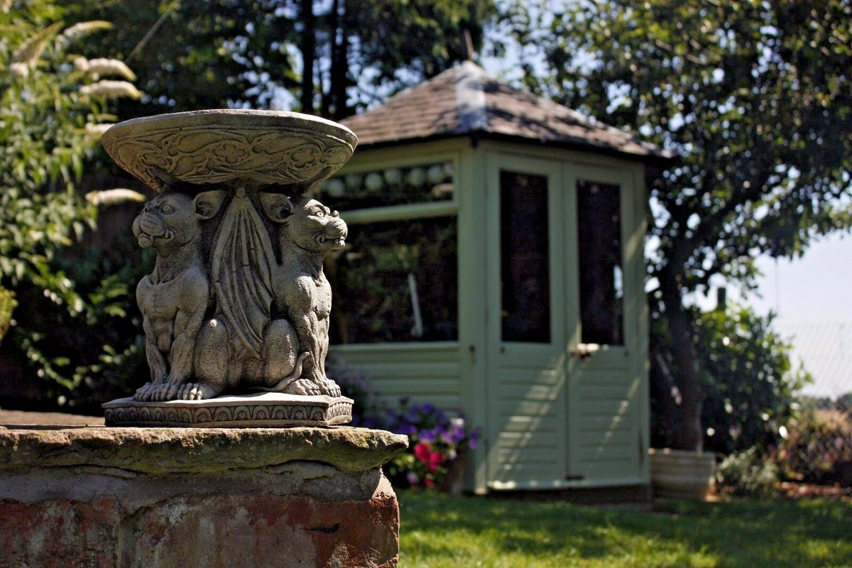 Stone Gargoyle Garden Bird Bath on a brick wall with a garden shed in the background, enhancing outdoor decor with gothic charm.
