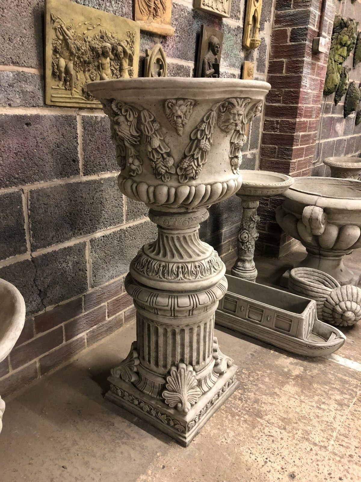 Single Bacchus design stone urn planter on ornate plinth, highlighting intricate carvings ideal for garden or patio decor.
