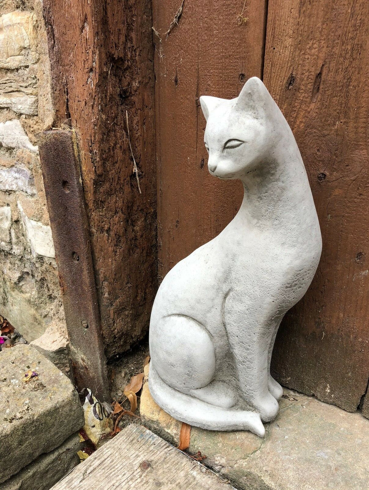 Single stone Siamese cat statue placed near a rustic corner, blending naturally with outdoor garden elements.