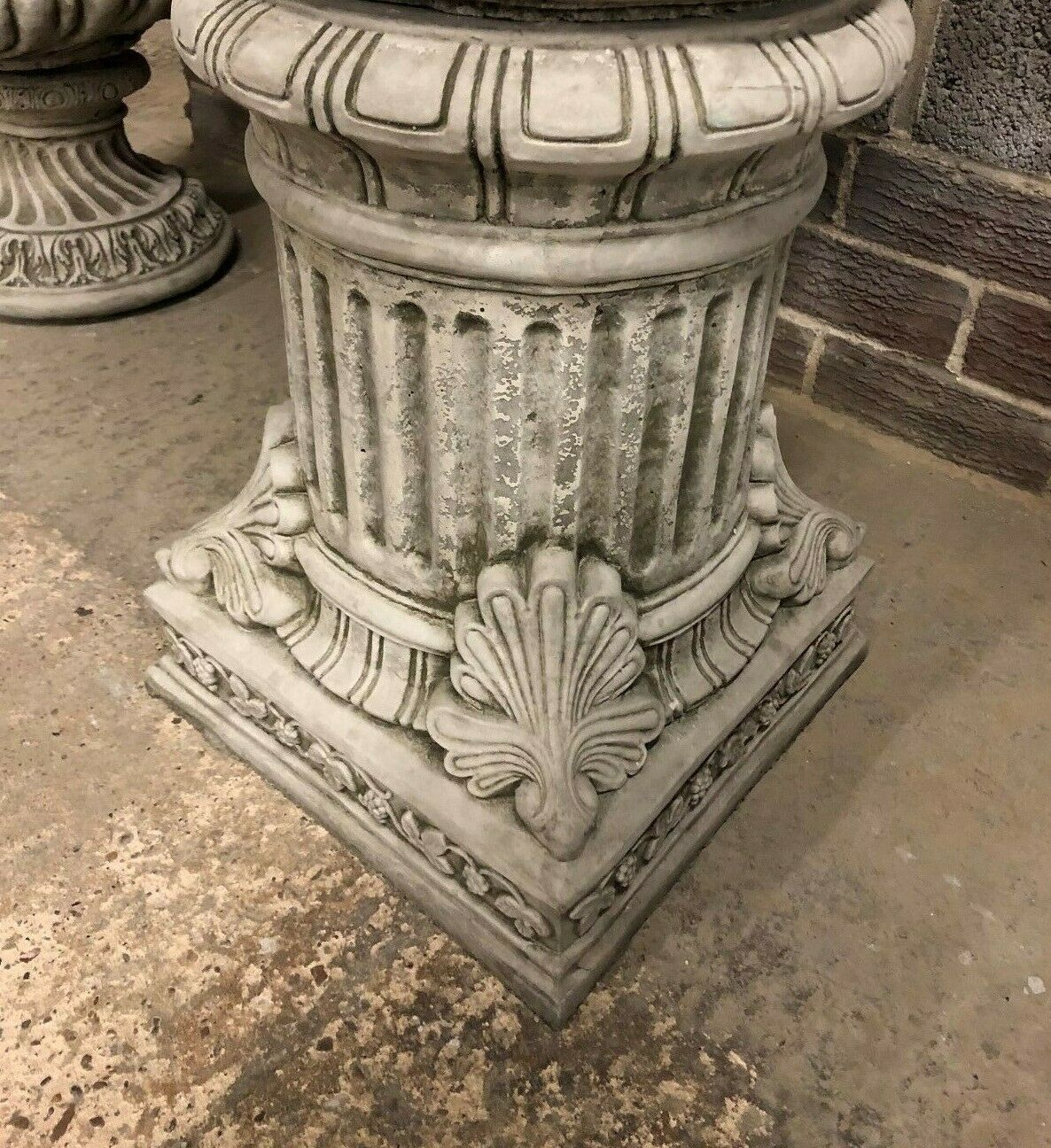 Detailed view of the base of a classical stone plinth with shell and floral motifs, suitable for supporting statues or planters in outdoor spaces.
